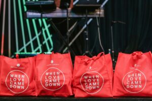 Red tote bags with “Victory Church” logo and “When Love Came Down” printed on them, displayed in front of a stage with musical equipment.